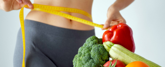 Desinchá mulher utilizando uma fita metrica na cintura perto de vegetais para começar uma dieta