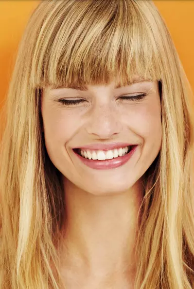 Mulher com franja e cabelos loiros sorrindo radiante com fundo laranja