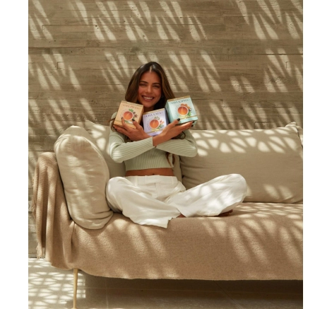 Kim Zucatelli setada em sofá marrom segurando três caixas de chá ayurveda