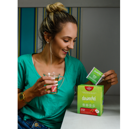 Mulher tomando chá de maçã com canela e segurando uma embalagem de Desinchá Sabores